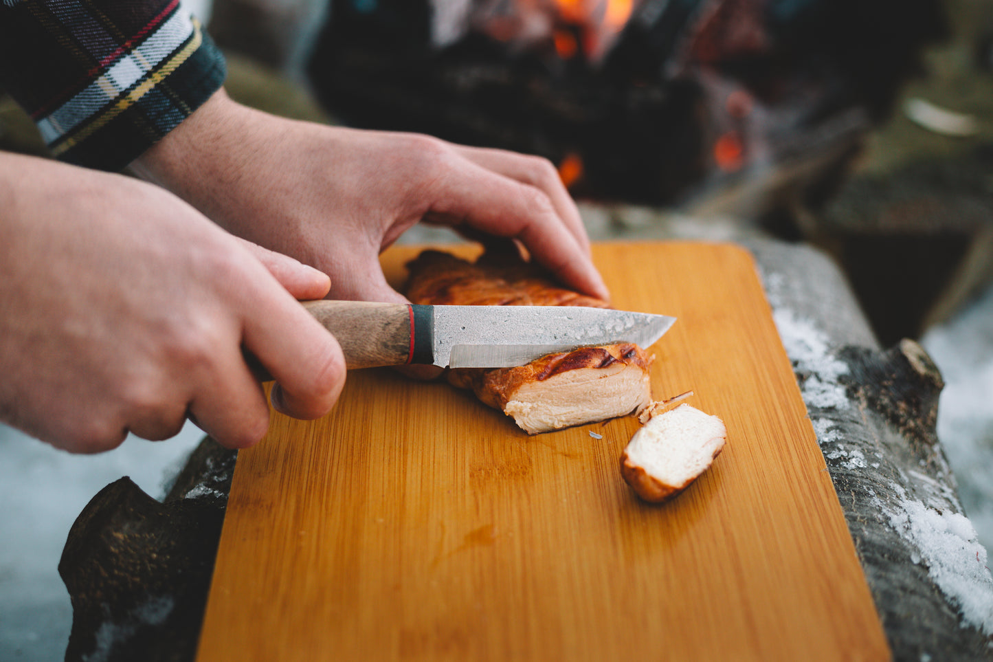 Person cutting bread on a wooden board with a knife, outdoor setting