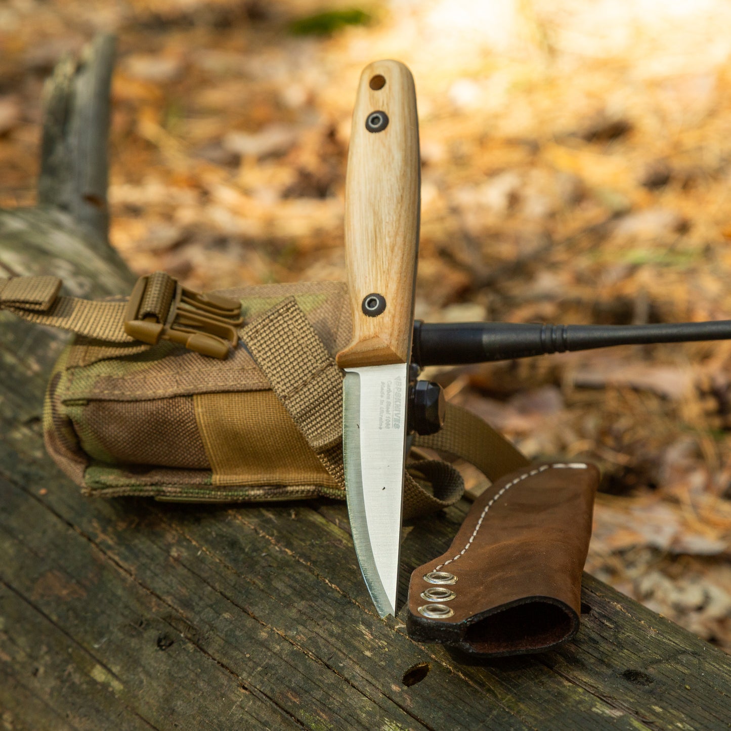 A fixed-blade knife with a light wood handle and silver blade is displayed upright on a weathered log outdoors, with a brown leather sheath and tan tactical pouch in the background.