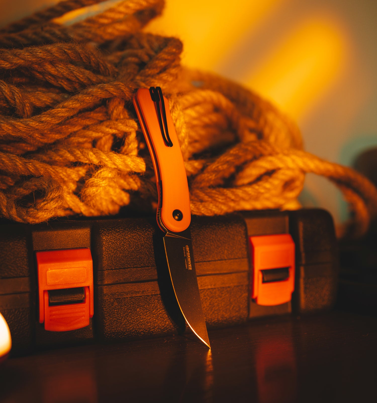 Orange pocket knife on a black sheath with red accents, next to coiled rope.