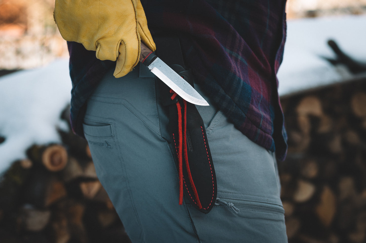 Person wearing yellow gloves holding a knife with a sheath against a blurred background of logs.