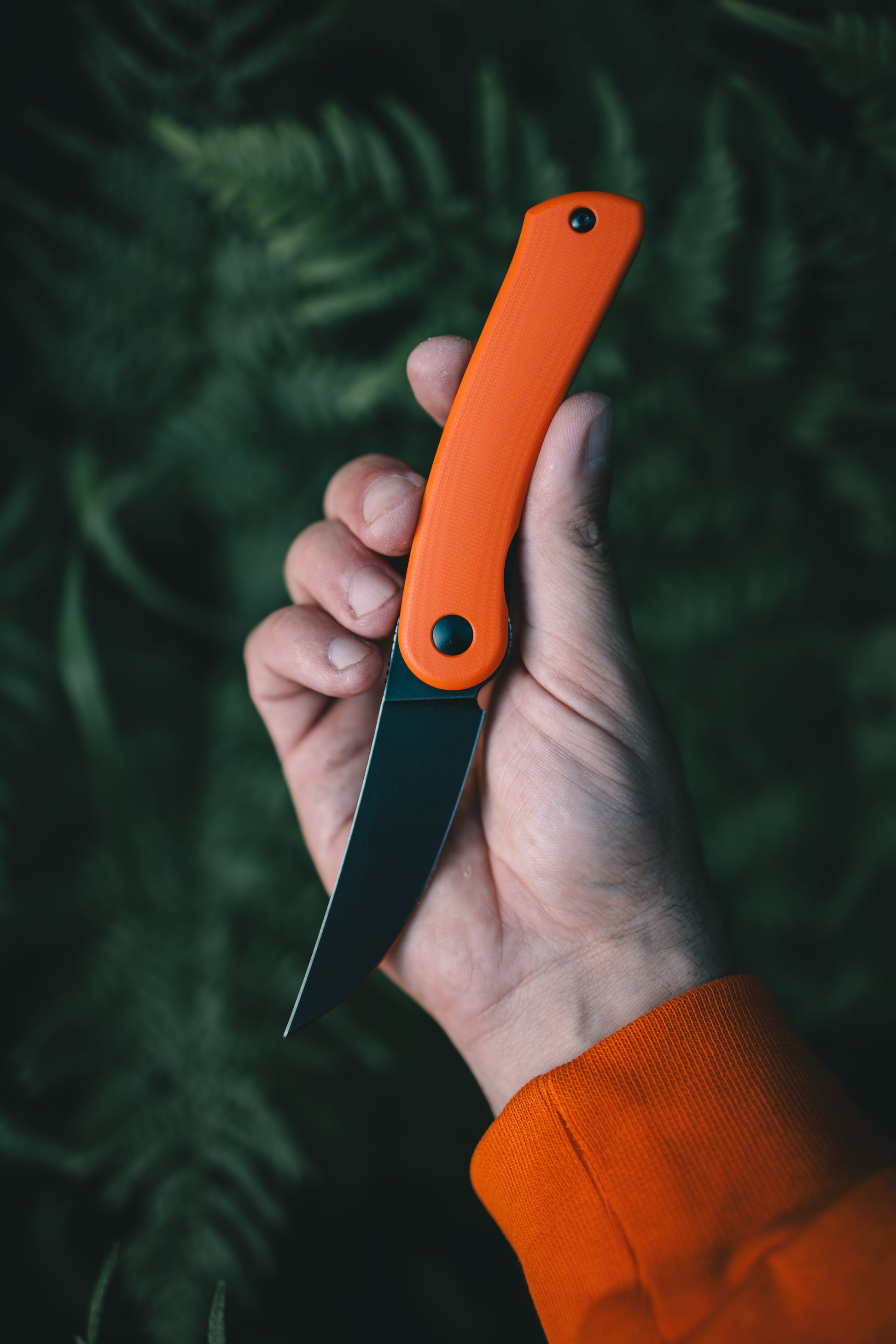 Hand holding a knife with an orange handle against a dark green leafy background