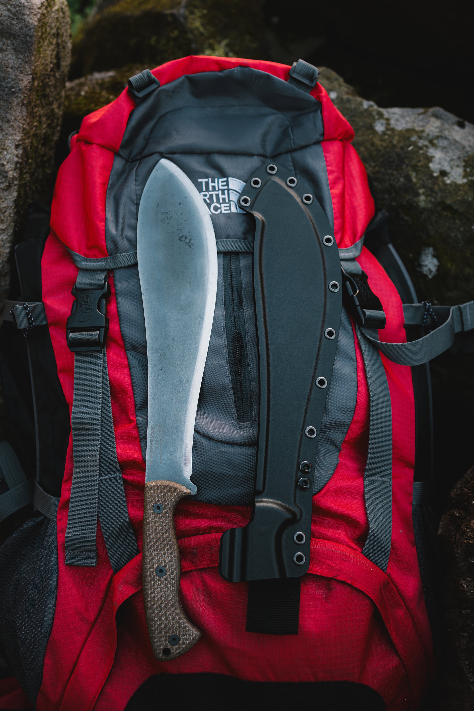 Red backpack with a machete attached, set against a natural rock background