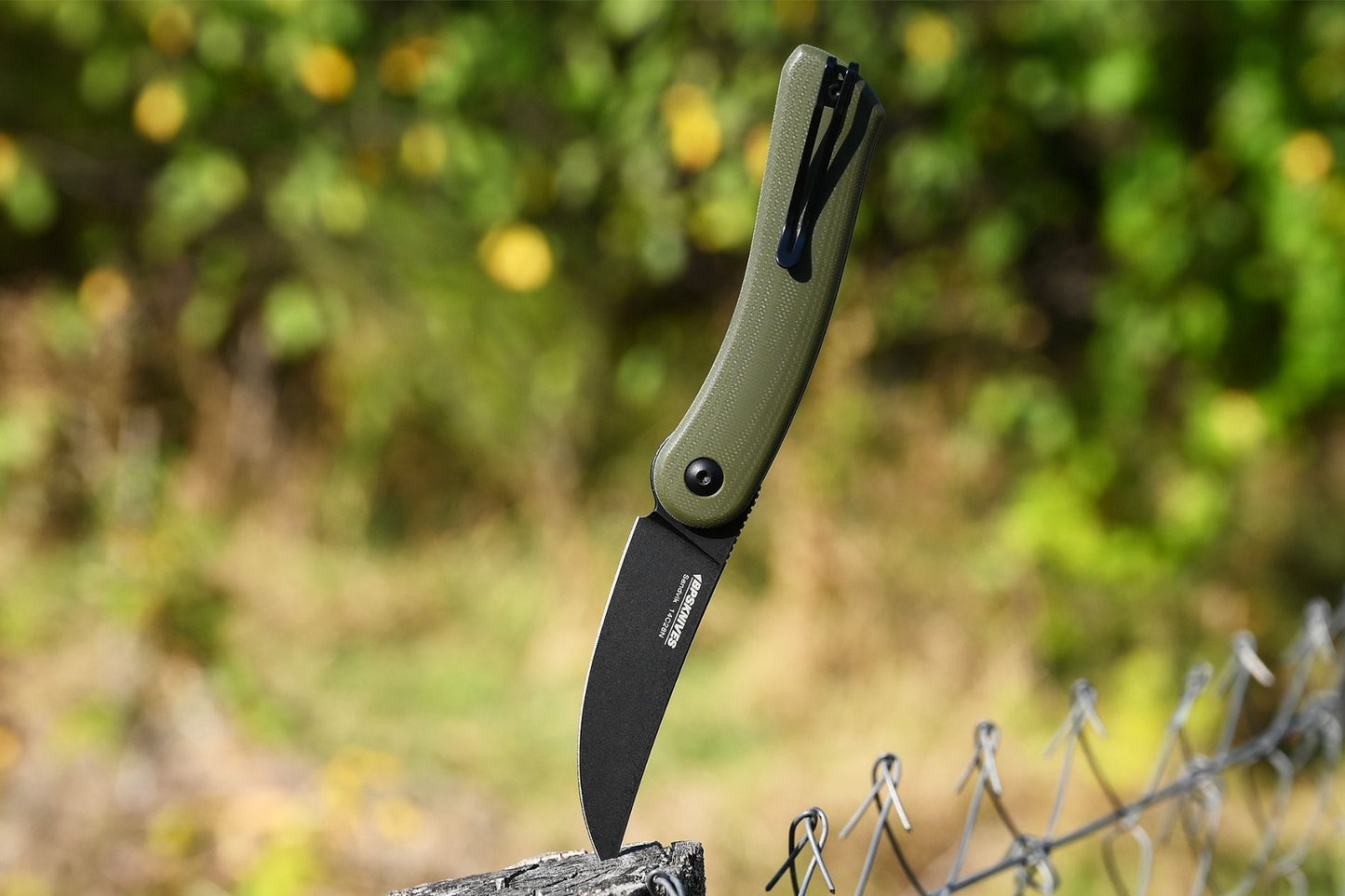 Folding knife with green handle and black blade on a wooden post against a blurred natural background