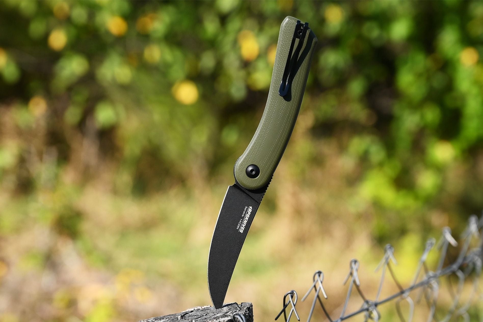 Folding knife with green handle and black blade on a wooden post against a blurred natural background