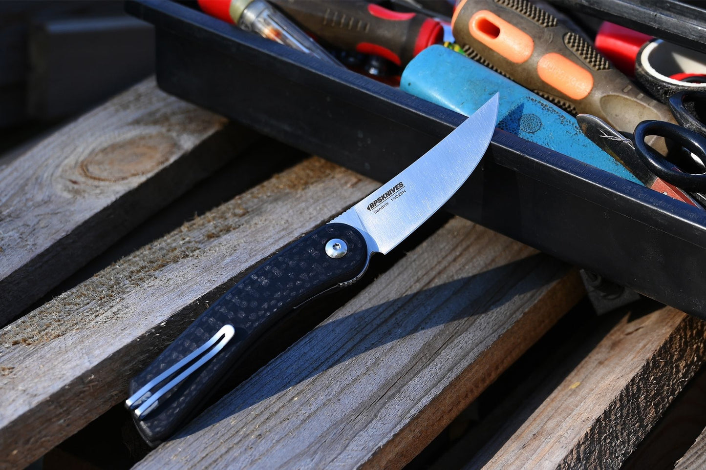 Folding knife with black handle and silver blade on a wooden surface with tools in the background