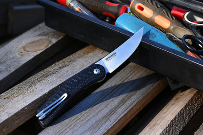 Folding knife with black handle and silver blade on a wooden surface with tools in the background