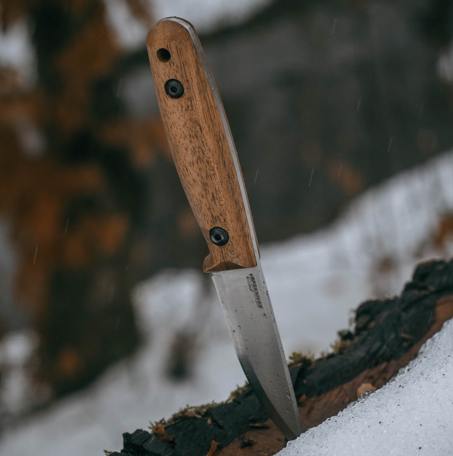 A fixed-blade knife with a brown wood handle and a silver steel blade, shown standing upright in a snowy outdoor setting.