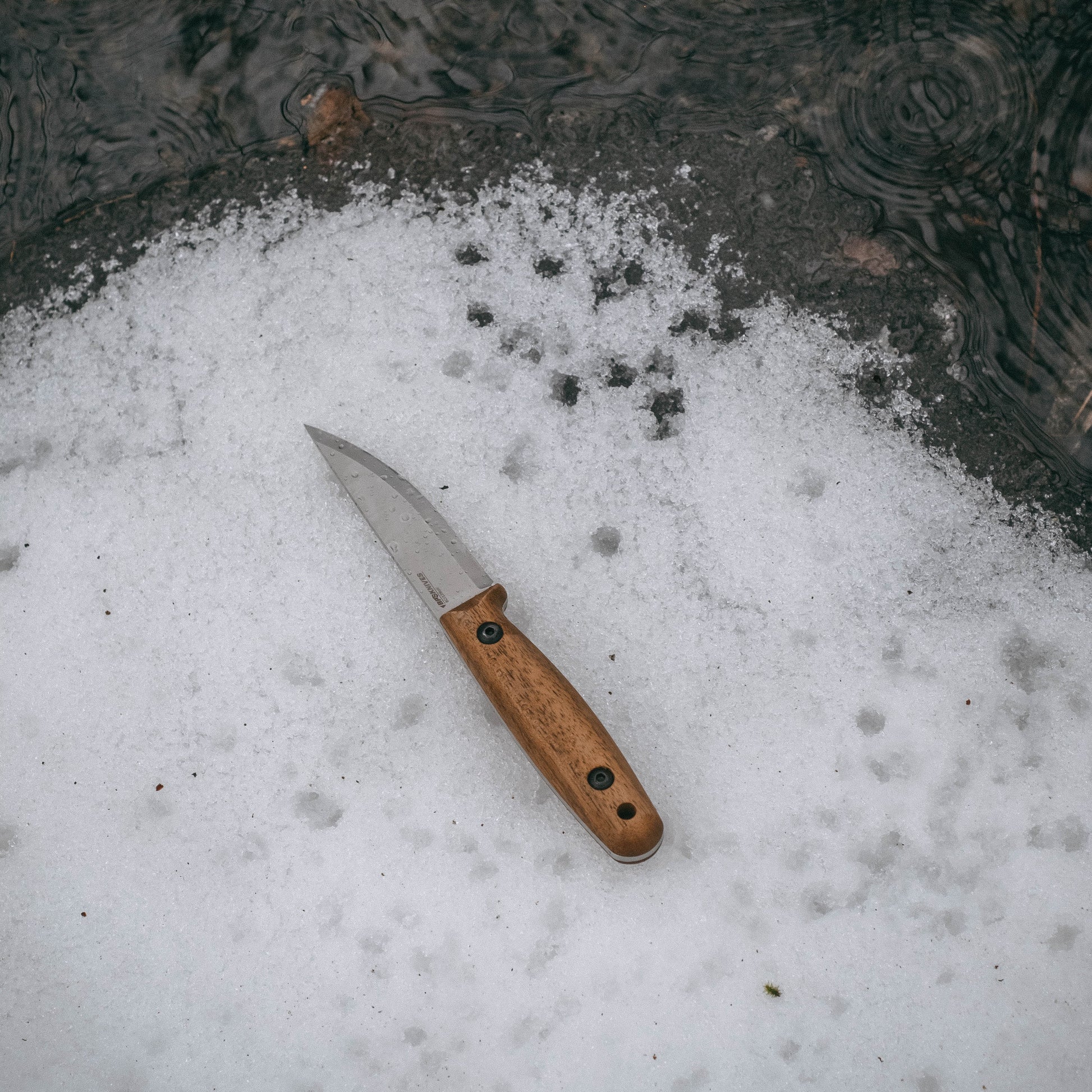 A fixed-blade knife with a silver steel blade and a light brown wooden handle lies on a patch of melting snow near dark water.