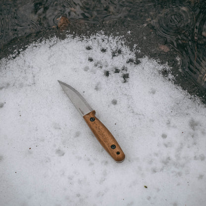 A fixed-blade knife with a silver steel blade and a light brown wooden handle lies on a patch of melting snow near dark water.