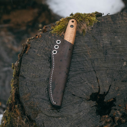 A fixed-blade knife with a light wood handle and a dark brown leather sheath, displayed on a mossy tree stump outdoors.