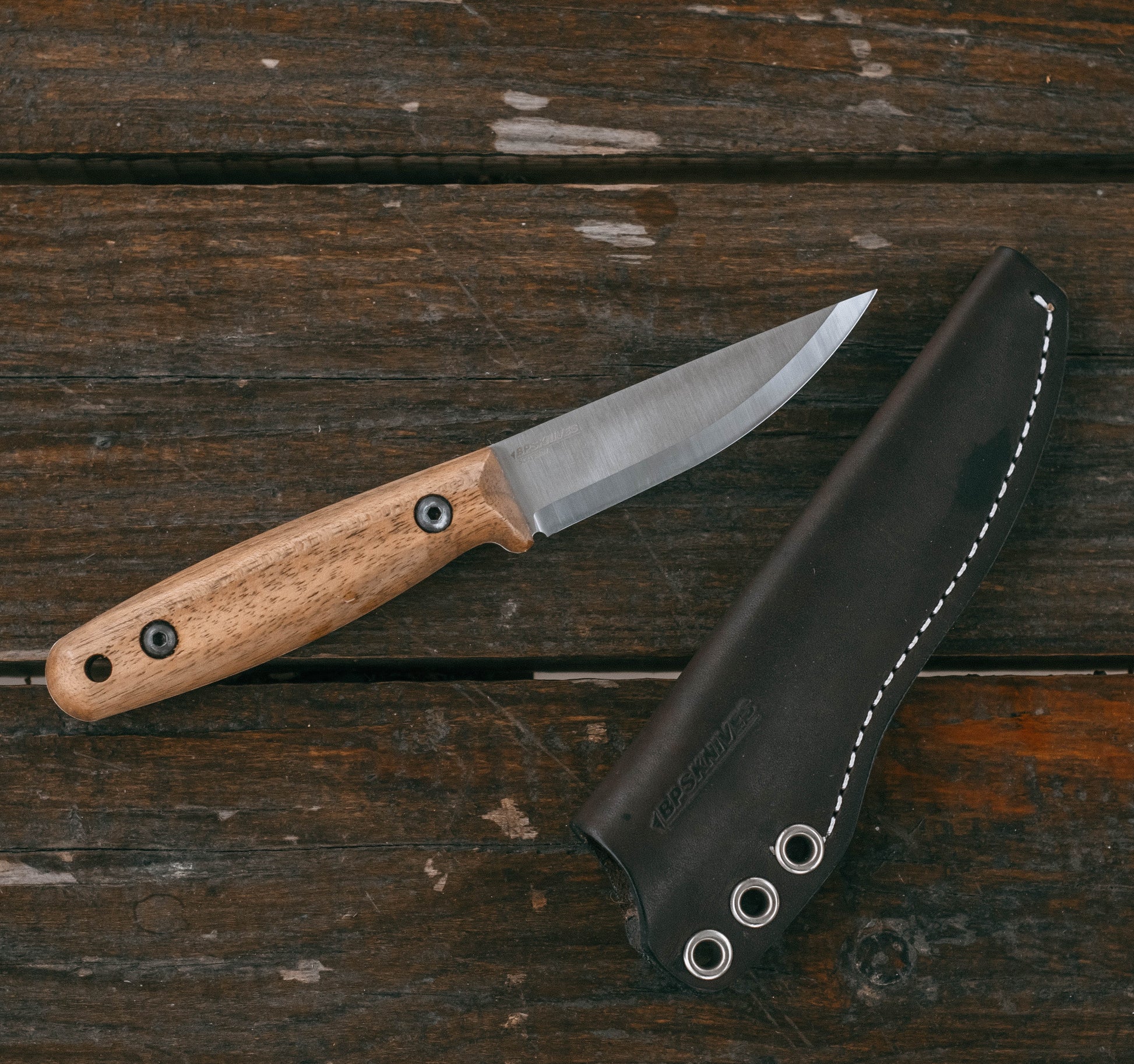 A fixed-blade knife with a light wood handle and satin-finished steel blade, placed diagonally next to its black leather sheath on a rustic dark wood plank surface.