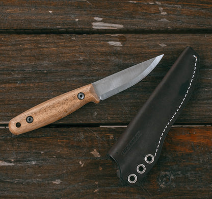 A fixed-blade knife with a light wood handle and satin-finished steel blade, placed diagonally next to its black leather sheath on a rustic dark wood plank surface.